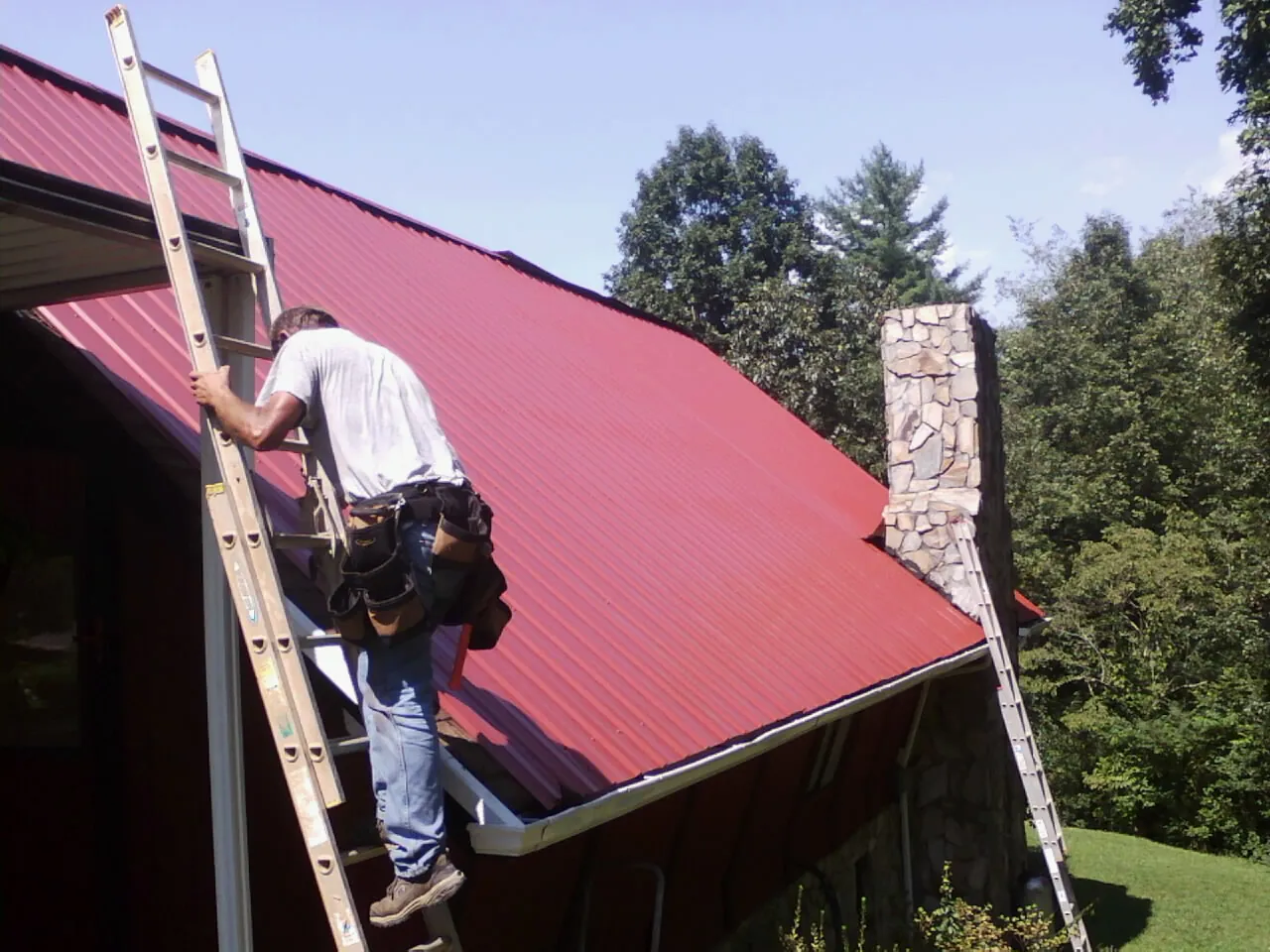 Roofer on a ladder beside a red metal roof and stone chimney — Soffit & Fascia Repair by Roofing Brothers in Hawarden